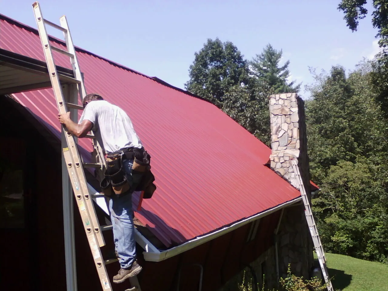 Roofer on a ladder beside a red metal roof and stone chimney — Soffit & Fascia Repair by Roofing Brothers in Watchung